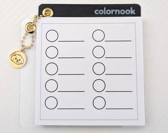 May include: A white notepad with a black top, featuring the text "colornook" in silver glitter. The notepad has ten circles with lines below them. A gold charm with a dog's face hangs from a gold chain.