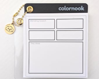 May include: A colour blending swatch book with the text "colornook" and a gold charm with a dog illustration. The swatch book has a black top and a clear plastic sleeve. The book has areas labelled "Blending Swatch" and "Test Area".