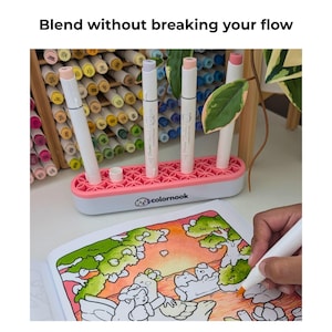 May include: A white and pink marker organizer with a floral-patterned silicone top, holding three white markers. The base features the text 'colornook' with a dog logo. A hand uses an orange marker to color a page in an open coloring book.
