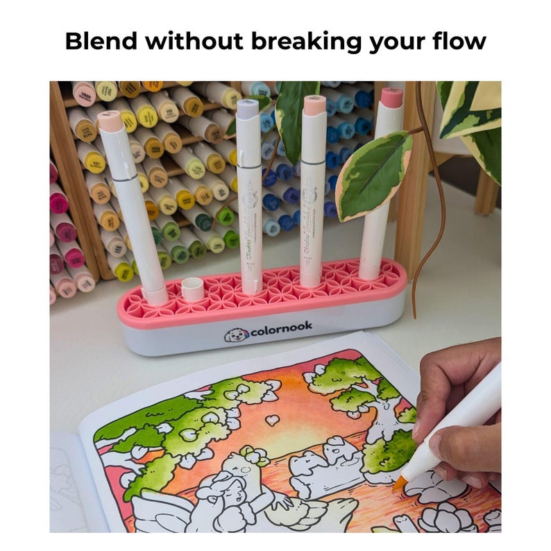 May include: A white and pink marker organizer with a floral-patterned silicone top, holding three white markers. The base features the text 'colornook' with a dog logo. A hand uses an orange marker to color a page in an open coloring book.