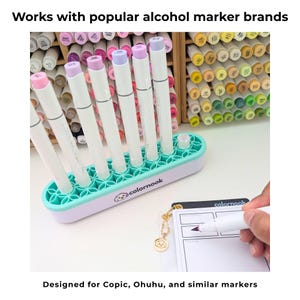 May include: A mint green and white oval marker organizer with a floral grid pattern, holding six white alcohol markers with purple caps. The base features the text colornook and a small dog logo. The text reads Works with popular alcohol marker brands.