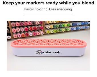 May include: A white rectangular marker organiser with a coral pink silicone top featuring a repeating geometric flower pattern. The front of the organiser displays the text colornook alongside a small graphic of a white dog with a blue ear.