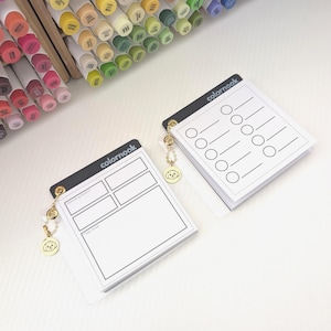 以下が含まれることがあります： 「colornook」の文字が入った黒いアクセントの白いノートパッドが2つ。1つのノートパッドには見本をテストするためのレイアウトがあり、もう1つには円と線があります。各ノートパッドには犬のデザインのチャームが付いています。背景にはカラフルなマーカーのコレクションがあります。