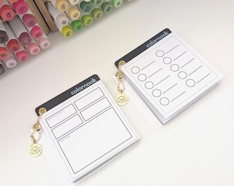 May include: Two white notepads with black accents and the text "colornook". One notepad has a layout for testing swatches, the other has circles and lines. Each notepad has a charm with a dog design. A collection of colourful markers is in the background.