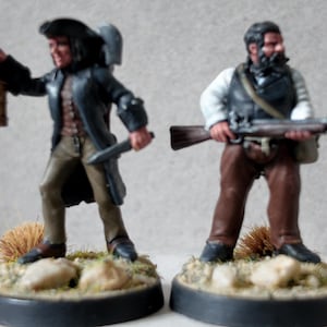 Puede incluir: Dos figuras en miniatura de personajes históricos. Una figura sostiene una linterna y un cuchillo, vistiendo un abrigo y sombrero negros. La otra figura sostiene un rifle, vestida con un chaleco y camisa blanca. Ambas figuras están sobre bases circulares.