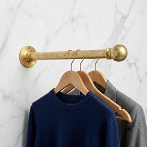 Puede incluir: Un perchero de pared de latón con una barra cilíndrica, un remate esférico y una placa de montaje circular. Tres perchas de madera cuelgan de la barra, sosteniendo un suéter azul marino y una chaqueta tipo blazer de color gris.