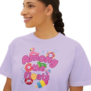 May include: A light purple t-shirt featuring a graphic print with the text Among Othe Floats in large pink letters. The design includes illustrations of a pink flamingo, a green turtle, colorful pool rings, a sun, and a popsicle surrounded by bubbles.