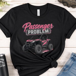 May include: Black t-shirt featuring a pink and black off-road vehicle graphic with the text "Passenger Problem" in pink script. The shirt is made of a soft material and has a crew neck.