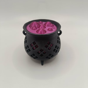 May include: A black three-legged cauldron with a lattice-patterned exterior and two small side handles. The top opening is filled with a flat, magenta-coloured insert featuring several raised, rounded, bubble-like protrusions of varying sizes.