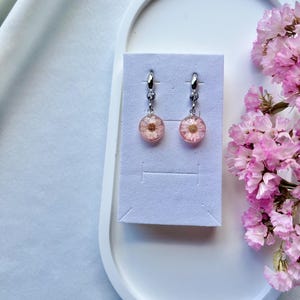 May include: A pair of silver-toned drop earrings featuring circular resin pendants. Each pendant contains a small, preserved pink daisy flower with a golden center. The earrings are attached to a light gray display card.