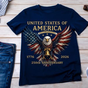 Op de afbeelding: Een marineblauw t-shirt met een afbeelding van een Amerikaanse zeearend met gespreide vleugels in de kleuren van de Amerikaanse vlag. De adelaar draagt een gouden ketting met een Liberty Bell. De tekst luidt: UNITED STATES OF AMERICA, LIBERTY'S SEMI-QUINCENTENNIAL, 1776, 2026, 250th ANNIVERSARY.