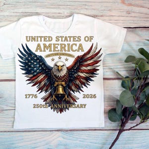 Op de afbeelding: Een wit t-shirt met een afbeelding van een Amerikaanse zeearend met gespreide vleugels in rood, wit en blauw. De adelaar draagt een gouden bel als ketting. De tekst luidt: UNITED STATES OF AMERICA, LIBERTY'S SEMI-QUINCENTENNIAL, 1776, 2026, 250th ANNIVERSARY.