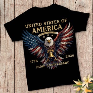 Op de afbeelding: Een zwart t-shirt met een afbeelding van een Amerikaanse zeearend met gespreide vleugels in rood, wit en blauw. De adelaar draagt een gouden ketting met een Liberty Bell-hanger. De tekst luidt: UNITED STATES OF AMERICA, LIBERTY'S SEMI-QUINCENTENNIAL, 1776, 2026, 250th ANNIVERSARY.