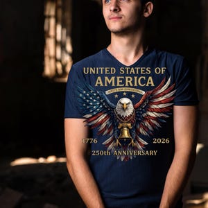 Op de afbeelding: Een marineblauw T-shirt met V-hals en een afbeelding van een Amerikaanse zeearend met gespreide vleugels in rood, wit en blauw. De tekst luidt UNITED STATES OF AMERICA, LIBERTY'S SEMI-QUINCENTENNIAL, 1776, 2026, 250th ANNIVERSARY, met een gouden bel.
