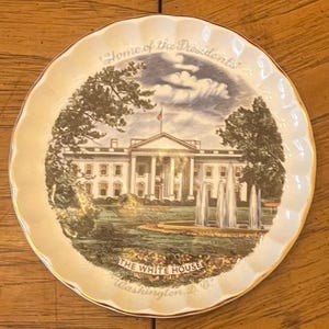 May include: A decorative plate with a scalloped edge and gold trim. The plate features an image of the White House, with the text "Home of the Presidents" and "The White House Washington D.C."
