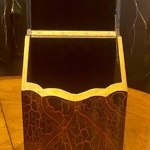 May include: A tall, upright wooden box with an open hinged lid. The exterior features a black and dark red crackle finish with a decorative scroll pattern at the base. The interior is lined with black fabric, and a metal hinge connects the lid.