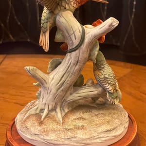 May include: A ceramic hummingbird sculpture featuring two hummingbirds perched on a tree branch. The birds have green, red, and brown coloring. The sculpture is mounted on a wooden base.