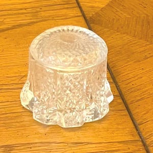 May include: A clear, cut-glass jar with a lid. The jar has a round, textured lid and a flared base. The glass has a diamond pattern. The jar is sitting on a wooden surface.