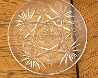 May include: A clear, round glass coaster with a starburst pattern. The coaster features a central star design surrounded by radiating star-shaped elements. The glass is transparent, allowing the wooden surface beneath to be visible.