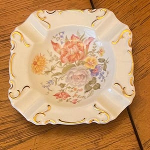 May include: Cream-colored ashtray with a floral design. The center features a bouquet of flowers in shades of orange, pink, yellow, and purple. The rim is edged with gold trim, and the ashtray has a square shape with rounded corners.