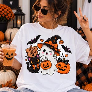 Peut inclure: Un t-shirt blanc avec un motif représentant un fantôme qui fait un clin d'œil avec un chapeau de sorcière, un chat noir avec un nœud orange, une citrouille, des chauves-souris et un panneau en bois indiquant TRICK OR TREAT.