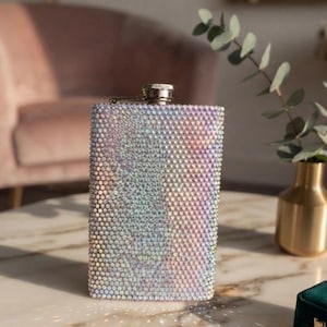 Op de afbeelding: Een rechthoekige metalen heupfles die volledig is bedekt met kleine, iriserende, lichtreflecterende kristallen die een glinsterend, veelkleurig effect creëren. De fles heeft een zilverkleurige schroefdop in het midden van de bovenrand.