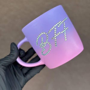 Pode incluir: Uma caneca degradê roxa e rosa com as letras "BFF" escritas em strass brilhantes. A caneca tem uma alça e é segurada por uma mão enluvada.
