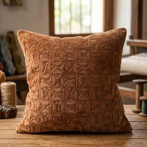 May include: A square throw cushion in a solid burnt orange colour featuring a repeating geometric pattern of interlocking circles and semi-circles embossed across the entire surface. The cushion has a soft, textured appearance and a plush, full shape.