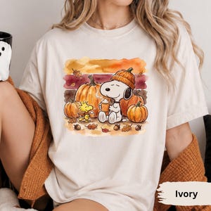 May include: An ivory t-shirt featuring a watercolor-style graphic of Snoopy wearing an orange knit hat and holding a steaming mug, sitting among pumpkins, acorns, and autumn leaves with Woodstock nearby. The design uses warm fall colors.
