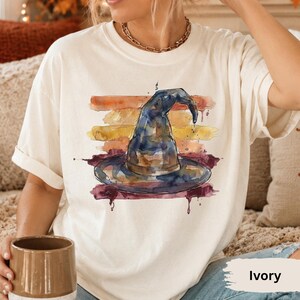 May include: An ivory short-sleeve t-shirt featuring a watercolor-style graphic of a dark blue witch hat centered over horizontal brushstrokes in shades of orange, yellow, and deep red. The design has a paint-splatter effect with visible drips.