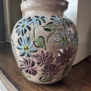 Puede incluir: Un jarrón de cerámica beige con acabado de esmalte craquelado y patrones florales recortados. Las flores están pintadas en tonos azul claro y morado oscuro con hojas verdes. El jarrón tiene un cuerpo ancho y redondeado con borde.
