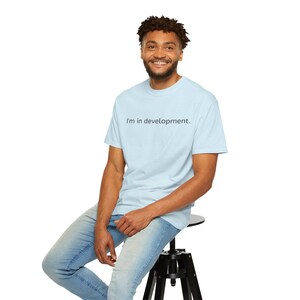 May include: A light blue t-shirt with the text "I'm in development." is worn by a person sitting on a black stool. The person is wearing blue jeans. The t-shirt is a solid color with a simple, minimalist design.