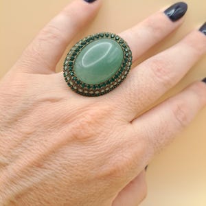 Può includere: Un anello di forma ovale con una grande pietra centrale liscia color verde chiaro. La pietra è circondata da un doppio bordo di piccoli cristalli sfaccettati verde scuro incastonati in una montatura in metallo scuro, creando un aspetto stratificato.