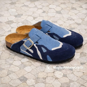 Könnte beinhalten: Ein Paar blaue Wildleder-Clogs mit Schnallenverschluss. Die Clogs haben ein zweifarbiges Design mit einer dunkelblauen Basis und hellblauen und weißen Akzenten. Das Fußbett ist hellbraun.