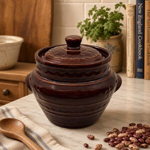 May include: A dark brown glazed ceramic bean pot with a matching lid, featuring a rounded body, horizontal ribbed detailing, and two small side handles. The pot has a scalloped edge design around the lid and the upper rim, with a glossy finish.