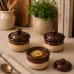 May include: Three round ceramic bowls with dark brown glazed lids and rims, featuring a light beige base. One bowl is filled with onion soup topped with a crouton. A matching brown lid sits beside it, and a metal spoon rests on a striped cloth.