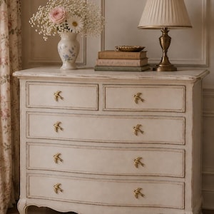 May include: Cream-colored dresser with six drawers, each adorned with gold bird-shaped handles. A vase of flowers and a lamp with a pleated shade sit atop the dresser. Books with gold lettering are stacked nearby, creating a vintage aesthetic.