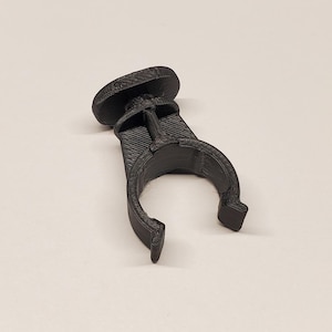 Könnte beinhalten: Eine schwarze Kunststoffklemme mit einem kreisförmigen Oberteil und einer gebogenen, U-förmigen Basis. Die Klemme hat eine strukturierte Oberfläche und ist zum Halten eines Objekts konzipiert. Das Objekt befindet sich vor einem hellen Hintergrund.