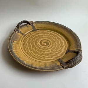May include: A round ceramic serving tray with two side handles, featuring a mustard yellow glaze with dark brown edges. The center of the tray displays a textured spiral pattern that radiates outward from the middle, creating a distinct visual effect.