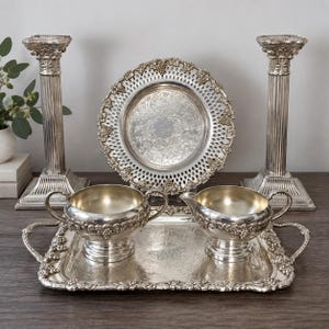 May include: A silver-toned tea service set featuring a rectangular tray with handles, a sugar bowl with two handles, and a creamer, all decorated with grape motifs. Behind the set are two tall fluted pillar candle holders and a round lattice plate.