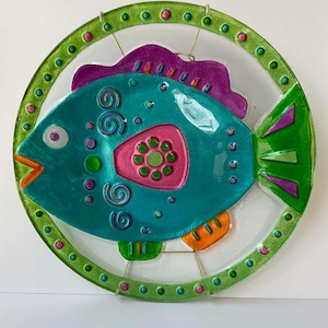 May include: A circular glass wall art piece featuring a teal fish with a purple dorsal fin, a pink side patch with green dots, and orange accents. The fish is framed by a lime green border decorated with small pink and teal dots throughout.
