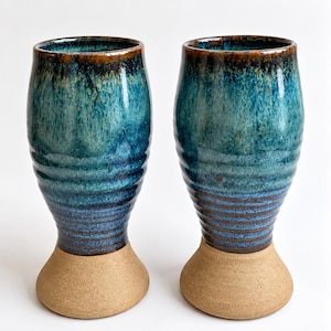 May include: Two tall ceramic tumblers featuring a flared base and a cylindrical body. The upper portion is glazed in a vibrant teal and blue drip pattern, while the lower section and base remain in an unglazed, textured tan clay finish.