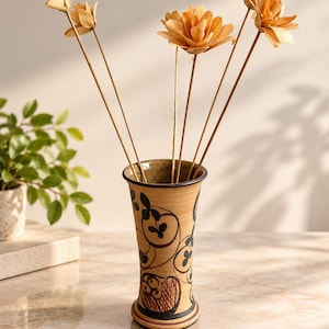 May include: A tall, flared ceramic vase with a tan base and dark brown hand-painted floral scrollwork and cross-hatched patterns. Five long, thin stems with light beige, layered wooden flower blossoms are arranged inside the vase.