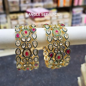 May include: A bangle set (6pcs) featuring gold-tone metal bangles adorned with teardrop-shaped clear stones, circular accents, and vibrant red and green enamel detailing. The intricate design includes repeating patterns of stones set in metal frames.
