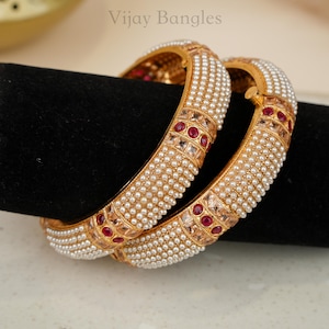 May include: Two gold-toned bangles featuring rows of small white pearls and square-cut clear stones accented by round red stones. The metal surface has a textured finish. The text Vijay Bangles is visible at the top of the image.
