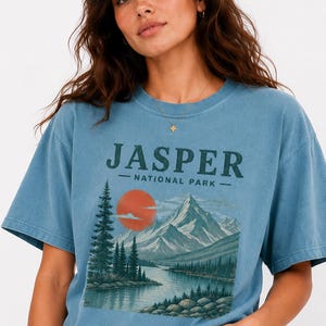 Pode incluir: Camiseta azul com o texto "JASPER NATIONAL PARK" acima de uma ilustra&ccedil;&atilde;o paisag&iacute;stica de montanhas, um lago e um p&ocirc;r do sol. A camiseta &eacute; de cor azul lisa e tem mangas curtas.