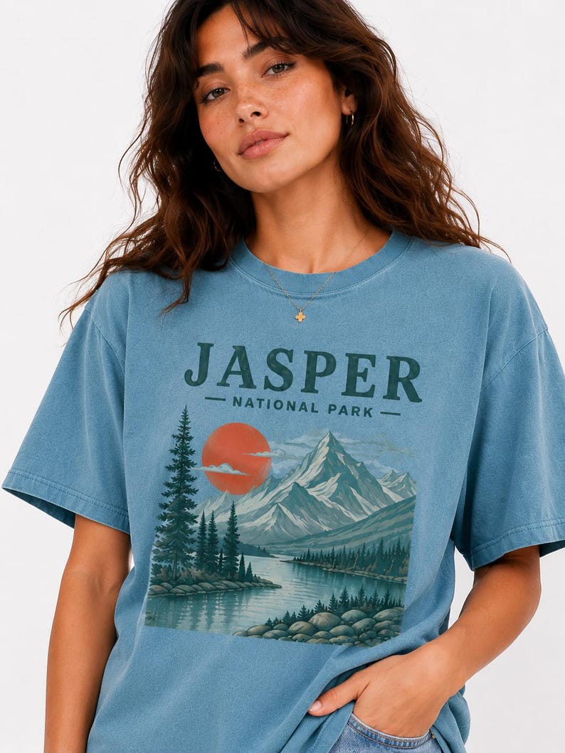 Pode incluir: Camiseta azul com o texto "JASPER NATIONAL PARK" acima de uma ilustra&ccedil;&atilde;o paisag&iacute;stica de montanhas, um lago e um p&ocirc;r do sol. A camiseta &eacute; de cor azul lisa e tem mangas curtas.