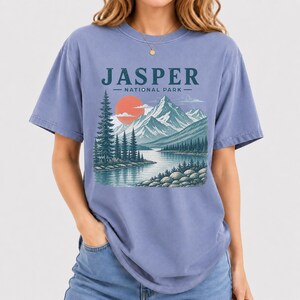 Pode incluir: Uma camiseta azul periwinkle com uma ilustra&ccedil;&atilde;o c&ecirc;nica do Parque Nacional Jasper. O design inclui um lago, montanhas, &aacute;rvores e um sol poente. O texto "JASPER - NATIONAL PARK" &eacute; impresso no topo.