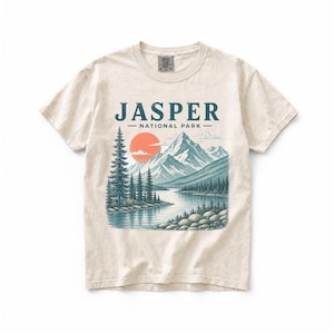 Pode incluir: Camiseta creme com uma ilustra&ccedil;&atilde;o c&eacute;nica do Parque Nacional Jasper. O design inclui uma cordilheira, um lago, &aacute;rvores e um sol poente. O texto "JASPER - NATIONAL PARK" est&aacute; impresso na parte superior.