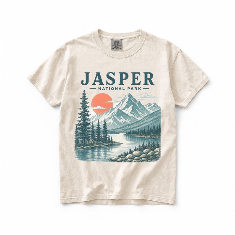 Pode incluir: Camiseta creme com uma ilustra&ccedil;&atilde;o c&eacute;nica do Parque Nacional Jasper. O design inclui uma cordilheira, um lago, &aacute;rvores e um sol poente. O texto "JASPER - NATIONAL PARK" est&aacute; impresso na parte superior.